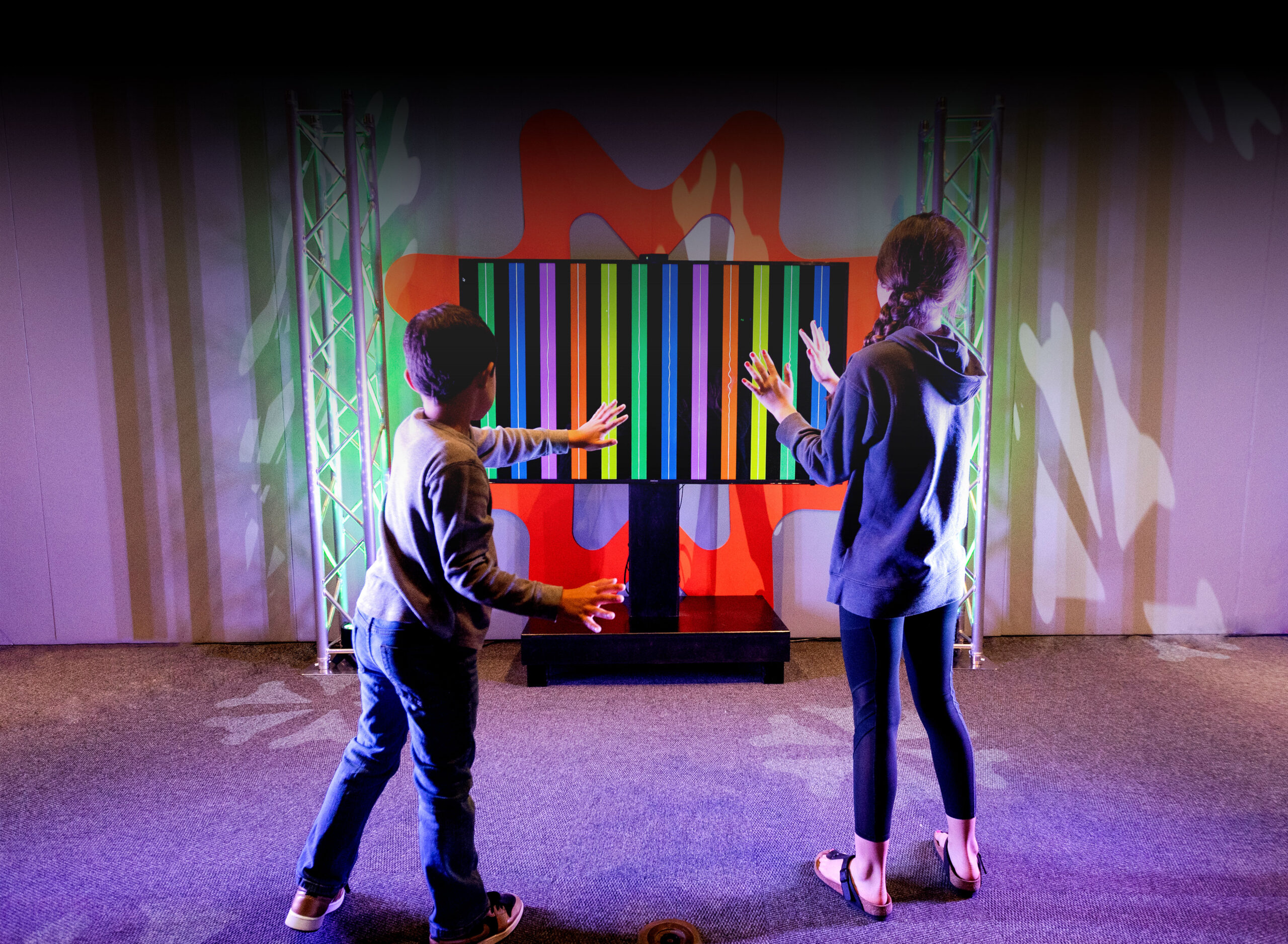 Two children waving their hands facing a monitor displayed colored soundwaves.