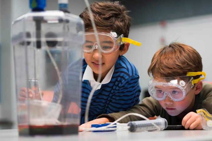 Two children wearing safety glasses are conducting a science experiment and looking at red liquid.
