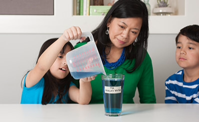 A child pouring liquid into a glass with an adult and another child watching.